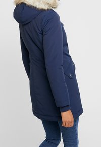 Navy blue parka jacket with a faux fur collar, zip closure, side pockets, and ribbed cuffs. Made from a smooth, insulated fabric.