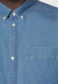 Camisa de mezclilla en azul claro con cuello abotonado, botones blancos y un bolsillo en el pecho. La tela tiene un acabado suave y ligeramente texturado.