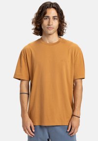Jonge man met schouderlang krullend haar draagt een losse bruine t-shirt en blauwe broek, staande tegen een effen lichte achtergrond.