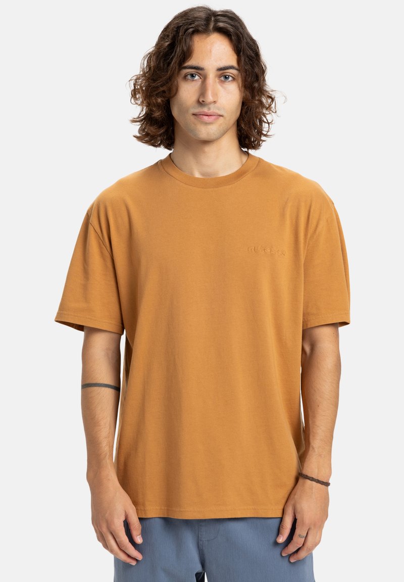 Jonge man met schouderlang krullend haar draagt een losse bruine t-shirt en blauwe broek, staande tegen een effen lichte achtergrond.