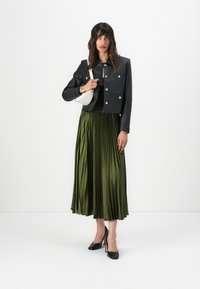 Veste cropped en cuir noir avec des boutons argentés, jupe plissée vert olive et escarpins noirs. Un sac à main blanc est porté sur l'épaule gauche.