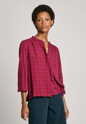 Blusa rossa a fantasia con bottoni sul davanti, tessuto morbido, rifinitura a volant sul collo e maniche a tre quarti, indossata con pantaloni scuri.