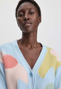 Hellblauer Cardigan mit V-Ausschnitt, der strukturierte pastellfarbene Patches in Rosa, Grün und Gelb aufweist. Er hat einen blauen Knopfverschluss.