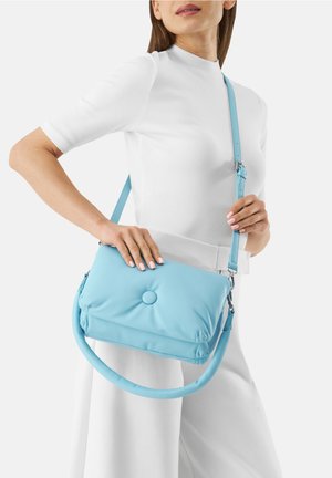 Žena oblečená v bílém krátkosrstém tričku a kalhotách drží světle modrou polstrovanou kabelku přes rameno s nastavitelným popruhem a ozdobným knoflíkem.