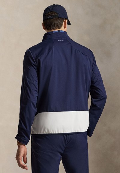 Muž nosící bundu s barevnými bloky v námořnické a bílé barvě a námořnickou čepici, zobrazen zezadu na jednobarevném béžovém pozadí.