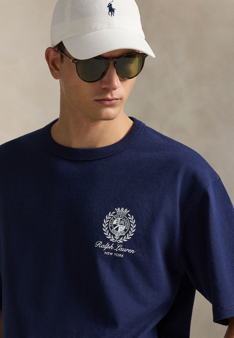 T-shirt en coton bleu marine avec un col ras du cou côtelé et un logo brodé blanc. Associé à une casquette blanche et des lunettes de soleil foncées.