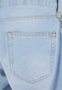 Világoskék farmeranyag, egyetlen hátsó zsebbel. A zseb sárga varrással készül a szélein, és sima, enyhén texturált felülettel rendelkezik.