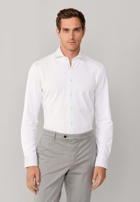 Mężczyzna w białej, długiej koszuli z guzikami włożonej w jasnoszare spodnie, stoi na tle jednolitego jasnoszarego tła.