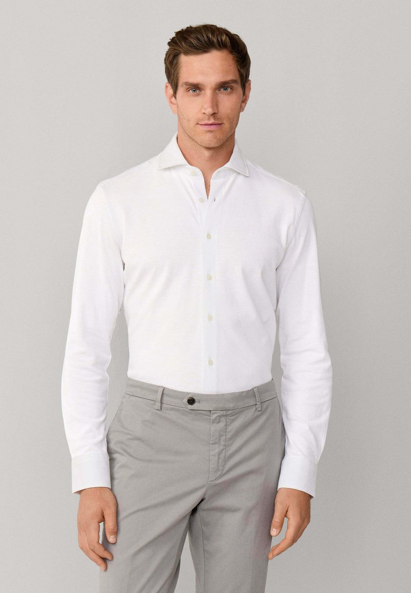 Mężczyzna w białej, długiej koszuli z guzikami włożonej w jasnoszare spodnie, stoi na tle jednolitego jasnoszarego tła.