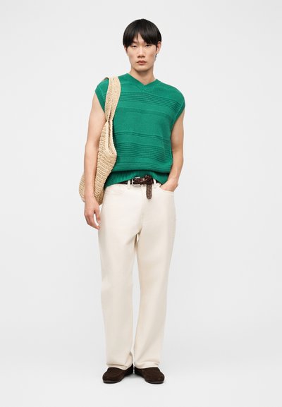 Jeune personne portant un haut vert sans manches en maille, un pantalon crème, des chaussures foncées, et un sac à bandoulière tissé beige, debout devant un fond uni.