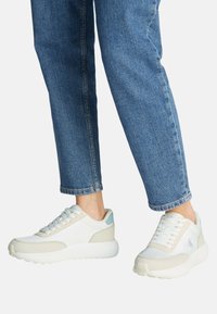 Zapatillas ligeras en blanco y beige, con un upper de textil suave, lengua acolchada y suela ligeramente texturizada. Acento sutil en azul.