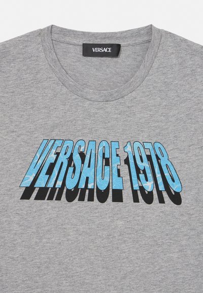 Camiseta gris de cuello redondo de Versace con texto azul "VERSACE 1978" y sombra negra centrada en el frente, mostrando la etiqueta Versace dentro del cuello.