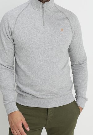 Mand iført en lysegrå quarter-zip pullover og olivengrønne bukser med den ene hånd i lommen.