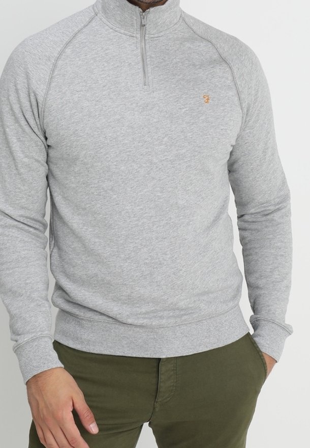 Homme portant un pull léger gris clair à fermeture éclair au quart et un pantalon vert olive, une main dans la poche.