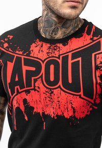 Camiseta de algodón negra con un bold logo rojo "TAPOUT" con patrón desgastado, cuello redondo y mangas cortas.