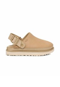 Mocassin en suède tan avec une sangle ajustable et une semelle en caoutchouc de couleur claire présentant un motif de semelle texturé. Marque UGG visible.