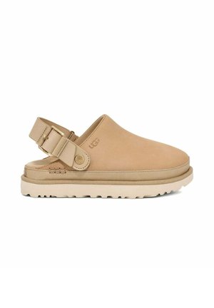 Zapato de ante marrón claro tipo slip-on con correa ajustable y suela de goma de color claro que presenta un patrón de textura en la banda de rodadura. Se ve el logo de UGG.