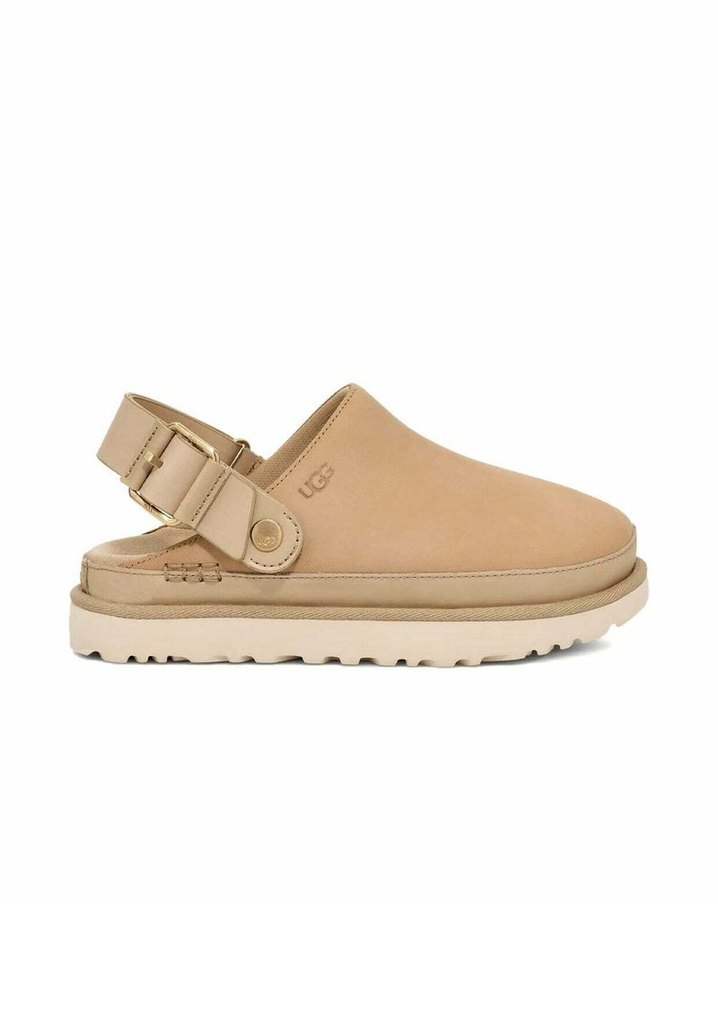 Mocassin en suède tan avec une sangle ajustable et une semelle en caoutchouc de couleur claire présentant un motif de semelle texturé. Marque UGG visible.