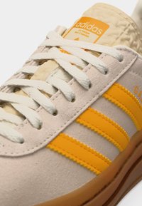 Μπεζ αθλητικό παπούτσι Adidas με λευκά κορδόνια, πορτοκαλί τρεις ρίγες, καουτσούκ σόλα και πορτοκαλί ετικέτα λογότυπου στη γλώσσα.