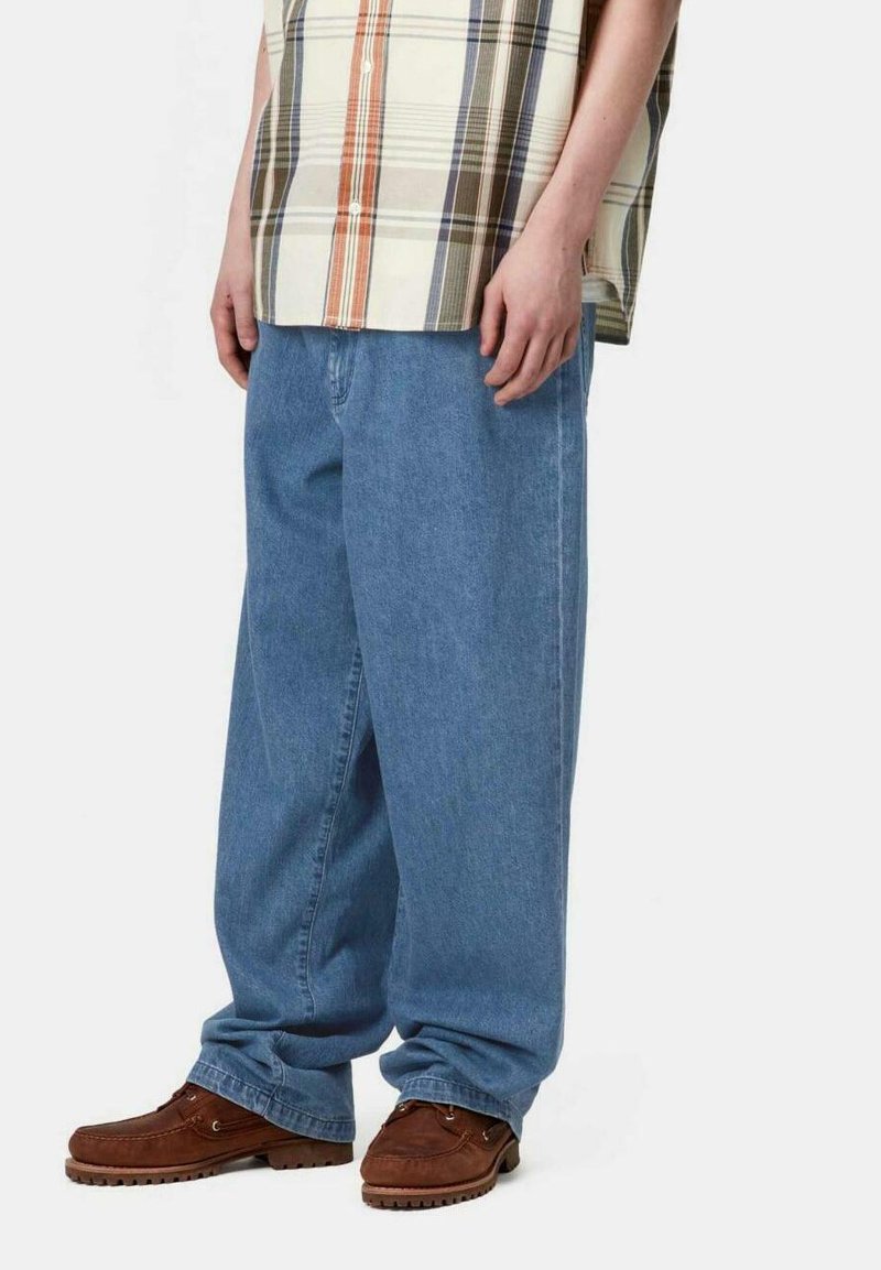 Jeans de mezclilla holgados de color azul claro, con textura suave, con bolsillos laterales, combinados con una camisa de cuadros multicolor de manga corta y mocasines marrones.