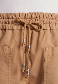 Tan shorts featuring an elastic waistband with a drawstring and silver bead accents, made of textured fabric with two side pockets.