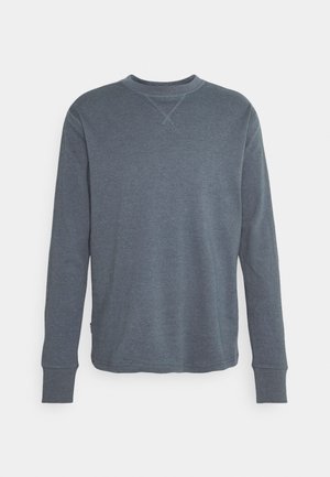 Mies, joka on pukeutunut pitkähihainen sininen paita, mustat kapealahkeiset housut ja mustat nahkasaappaat, seisoo valkoista taustaa vasten.
