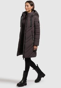 Donna che cammina indossando un lungo cappotto trapuntato marrone scuro con cappuccio, leggings neri e stivali neri fino al ginocchio su uno sfondo chiaro.