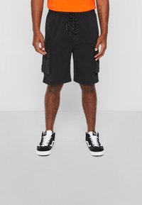 Shorts cargo noirs avec une taille à cordon, dotés de deux poches latérales. Accompagnés de baskets noir et blanc. Tissu lisse, coupe au genou.