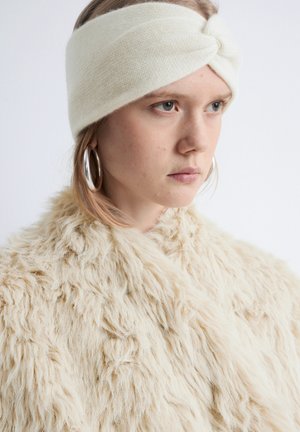 Cream knitted headband with a twisted design, worn with a beige, fluffy faux fur coat. The headband is smooth, and the coat has a textured finish.