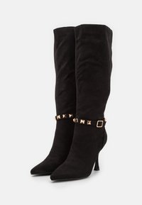 Botas de ante negras hasta la rodilla con puntera afilada, que presentan una correa en el tobillo adornada con tachuelas doradas y un delgado tacón de aguja. Textura suave.