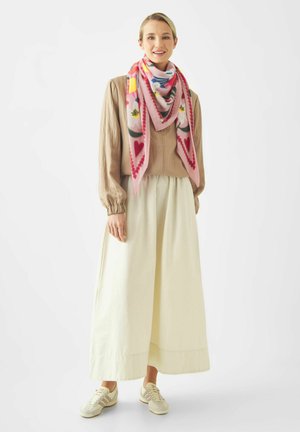 Jonge vrouw in beige blouse, lange witte rok, kleurrijke bloemen sjaal en witte sneakers, staand tegen een effen lichte achtergrond.