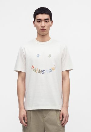 Jeune homme portant un T-shirt blanc avec un motif de texte coloré et courbé et un pantalon beige, debout devant un fond blanc uni.