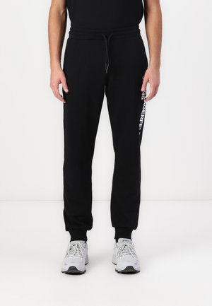 Black sweatpants in soft fabric with a drawstring waist, tapered legs, and a printed logo detail on the left side. Paired with gray sneakers.