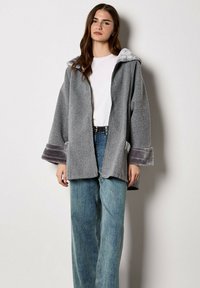 Giacca grigia oversize con cappuccio e polsini in morbida pelliccia sintetica. Presenta una chiusura con cerniera e due tasche frontali, abbinata a jeans azzurri.