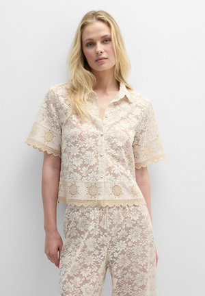 Mujer con cabello largo y rubio, lleva una camisa corta de manga corta con encaje crema y pantalones de encaje a juego, de pie frente a un fondo blanco liso.