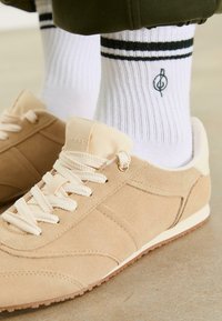 Persona che indossa sneakers beige in suede con spesse sciarpe bianche e calze bianche a costine con strisce nere e un logo circolare, in piedi su un pavimento.