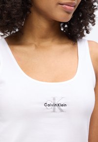 Γυναίκα με λευκό τοπ αμάνικο με το λογότυπο "Calvin Klein" σε μαύρο και γκρι, με σγουρά μαλλιά ορατά, κομμένη από τα χείλη μέχρι τον άνω κορμό.