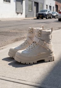 Chunky beige high-top boots with thick soles and straps labeled "Buffalo" placed on a sunlit urban sidewalk.