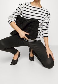 Black woven clutch bag held in front, featuring a textured surface. The user wears a white and black striped shirt and black pants.