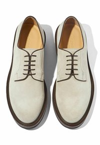 Scarpe in suede beige chiaro con punta arrotondata e dettagli in pizzo marrone. Presentano una suola marrone a contrasto e sottili accenti di cucitura.