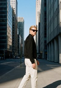 Homme blond portant des lunettes de soleil, une veste noire et un pantalon blanc marchant sur un trottoir ensoleillé de la ville entre de grands immeubles en verre et en béton.