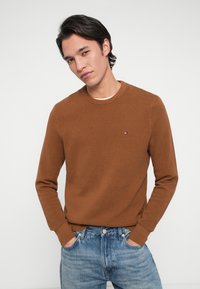 Brauner strukturierter Strickpullover mit Rundhalsausschnitt, versehen mit einem kleinen Logo auf der linken Brust, kombiniert mit hellblauen Jeans.