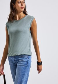 Jasnozielony, bezrękawnik o luźnym kroju i teksturowanej tkaninie, zestawiony z jasnoniebieskimi dżinsami i czarną bransoletką na nadgarstku.