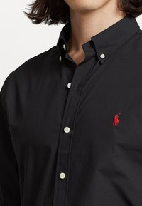 Svart knapp-up skjorta med klassisk krage, prydd med vita knappar och en liten röd broderad logotyp på bröstet. Slät tygtkänsla.