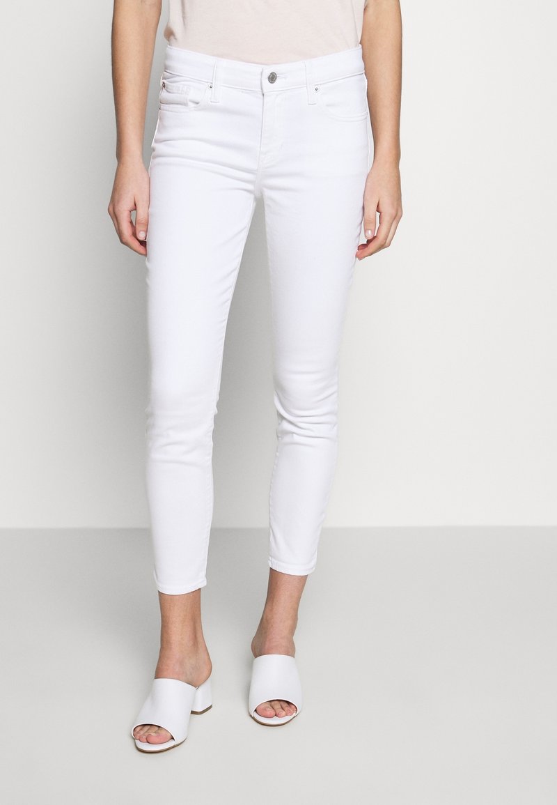 Femme portant un pantalon blanc ajusté et court ainsi que des mules blanches à talons ouvertes à l'avant, debout devant un fond clair uni.