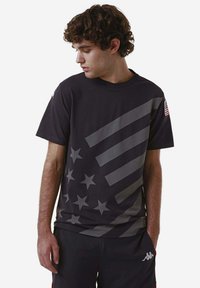 T-shirt nera in cotone con strisce diagonali grigie e motivi a stelle, colletto girocollo e un accento della bandiera americana sulla manica.