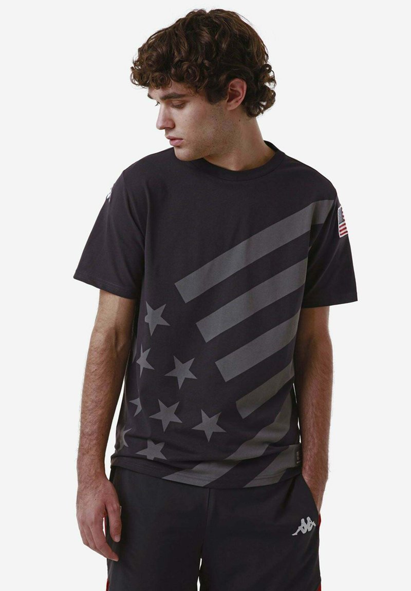 T-shirt nera in cotone con strisce diagonali grigie e motivi a stelle, colletto girocollo e un accento della bandiera americana sulla manica.