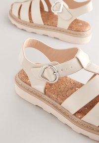 Sandales de couleur claire avec semelle intérieure en liège, larges brides et boucle en argent en forme de cœur. Semelle épaisse avec une finition texturée.