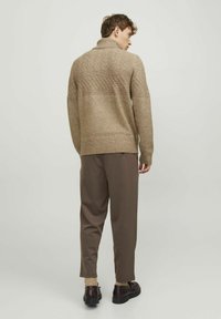 Maglione lavorato a maglia beige con colletto alto e motivo testurizzato, abbinato a pantaloni marroni e scarpe scure. L'aspetto è definito e rilassato.