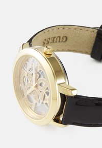 Orologio dorato con un quadrante trasparente che rivela dettagli intricati. Cinturino in pelle nera lucida, con accenti testurizzati e una corona sul lato.